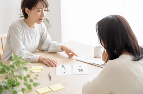 机の中央に「買い切り」「月額」と書かれた2枚のカードを並べ、2人の担当者が指差しながら向き不向きを話し合っている打ち合わせの一場面