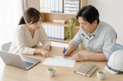 現場事務所の打ち合わせテーブルで、現場所長と事務担当が1枚の工事打合せ簿を一緒に覗き込みながら、日時欄と本文欄を確認して書き込んでいる場面