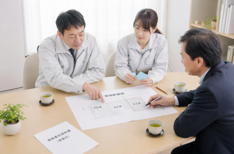 現場事務所の打合せテーブルで、現場所長・事務担当・外部担当の3人が現場配置図を囲み、C工区だけをマーカーで囲みながら現場単位の契約を打合せている風景